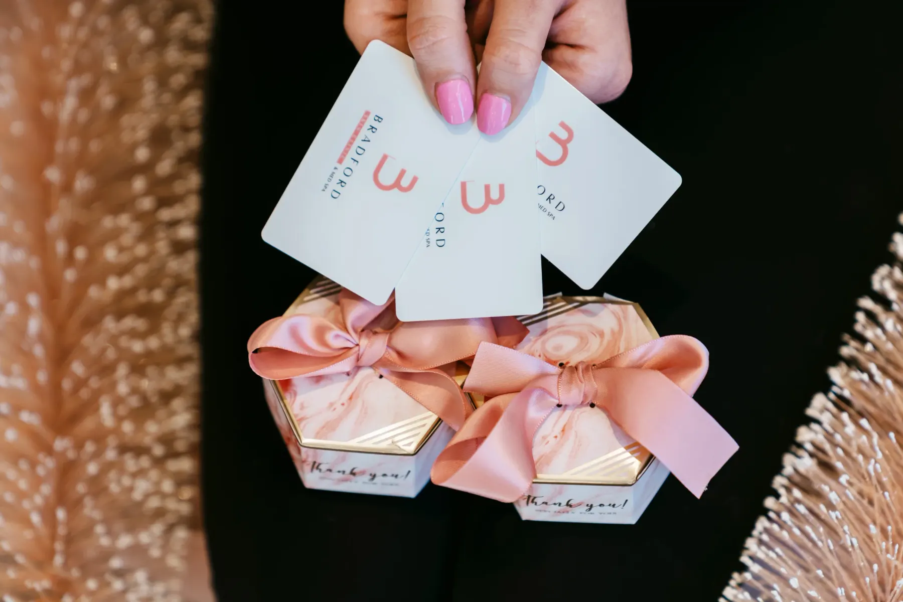 A hand holding three Bradford Skin Clinic gift cards above two small gift boxes tied with pink ribbon.