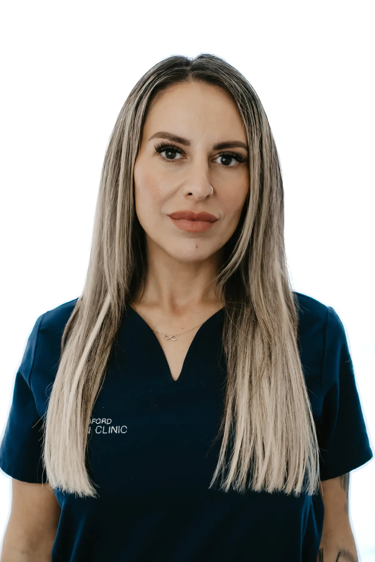 A member of the clinic team with long blonde hair, in Bradford Skin Clinic navy scrubs, against a pale background.