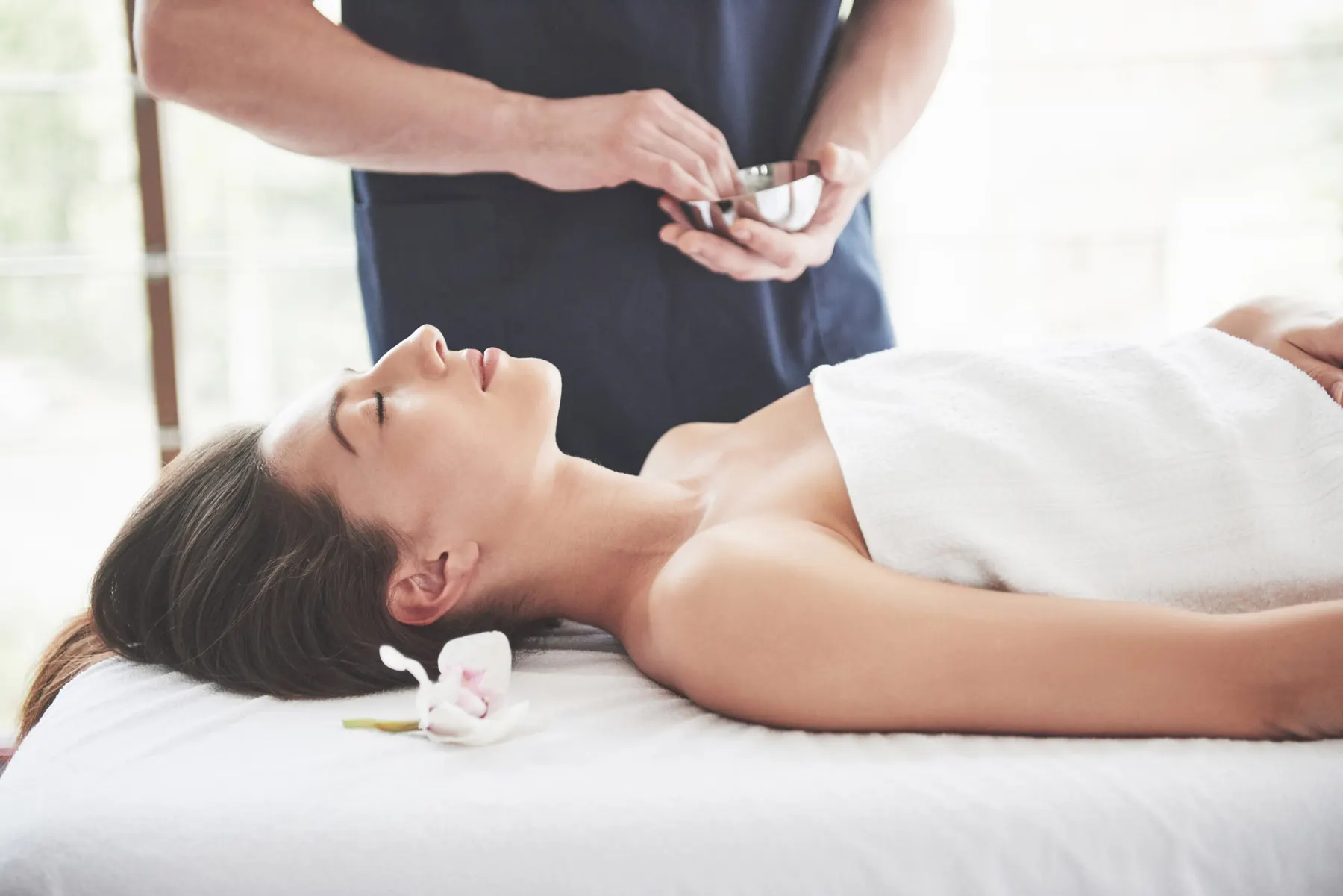 A client lying back on a treatment couch while a practitioner mixes product in a small bowl
