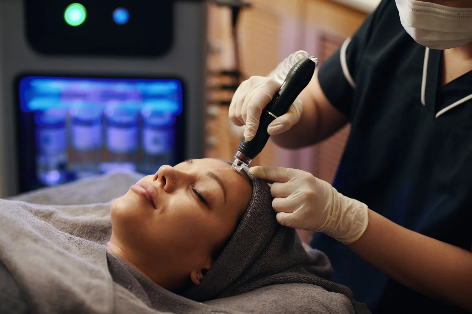 A client lying back with her eyes closed while a practitioner in white gloves runs a treatment pen across her forehead.