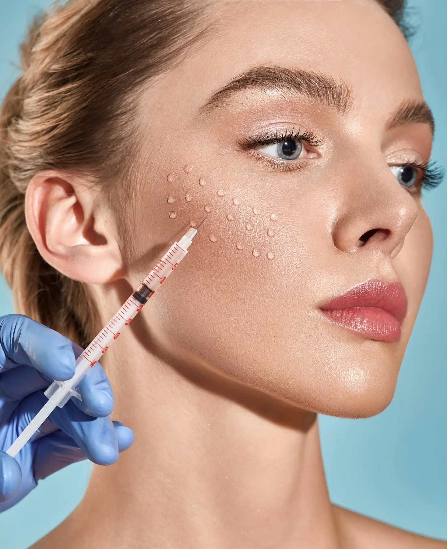 A woman against a pale blue background with a row of small marker dots on her cheek and a syringe held beside them.