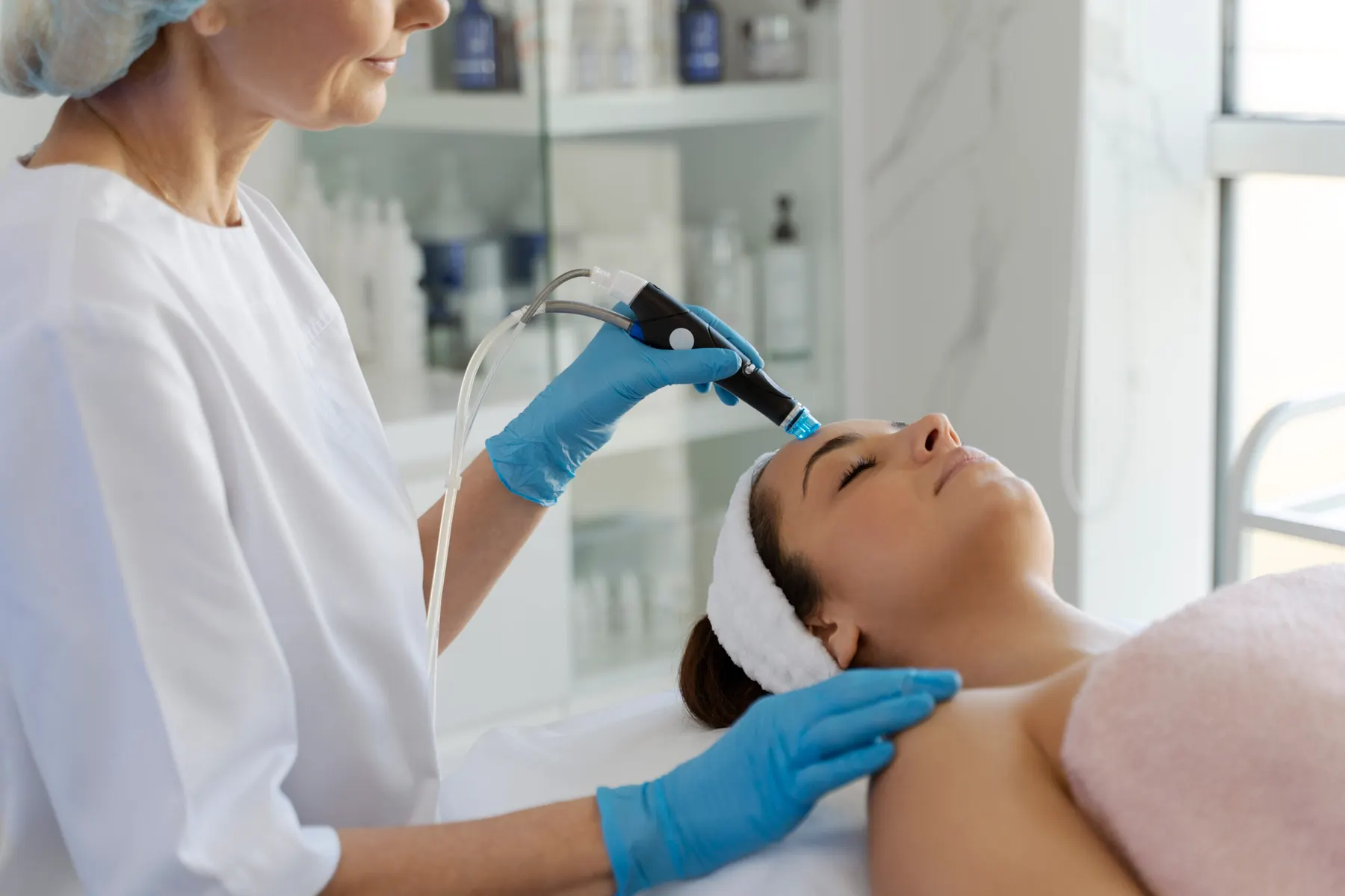 A practitioner in blue gloves guiding a treatment handpiece across the cheek of a client lying on a clinic bed.
