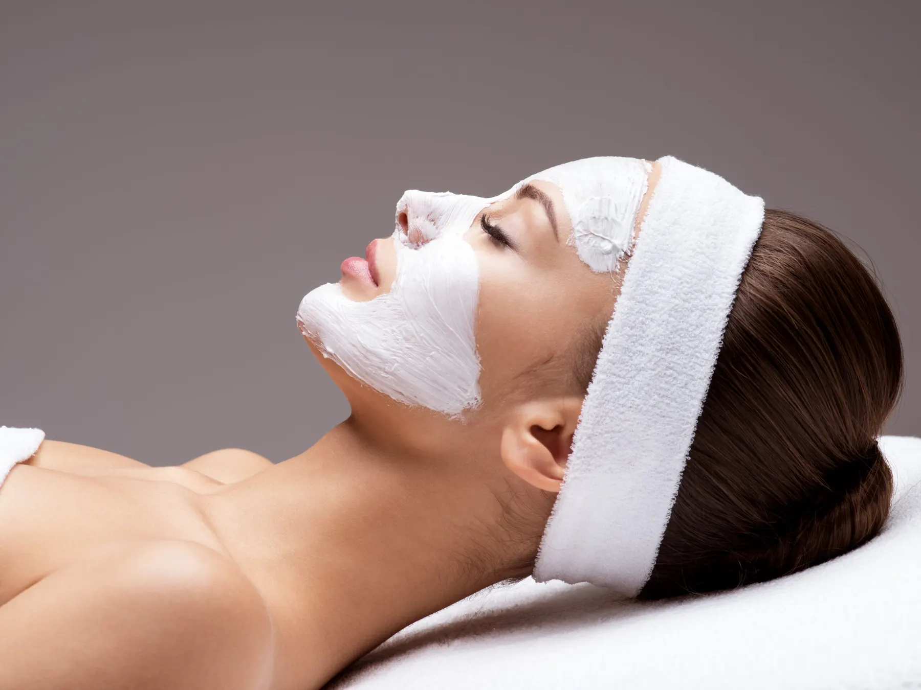 A client lying back with a white treatment mask applied over her face and a towel around her hair.