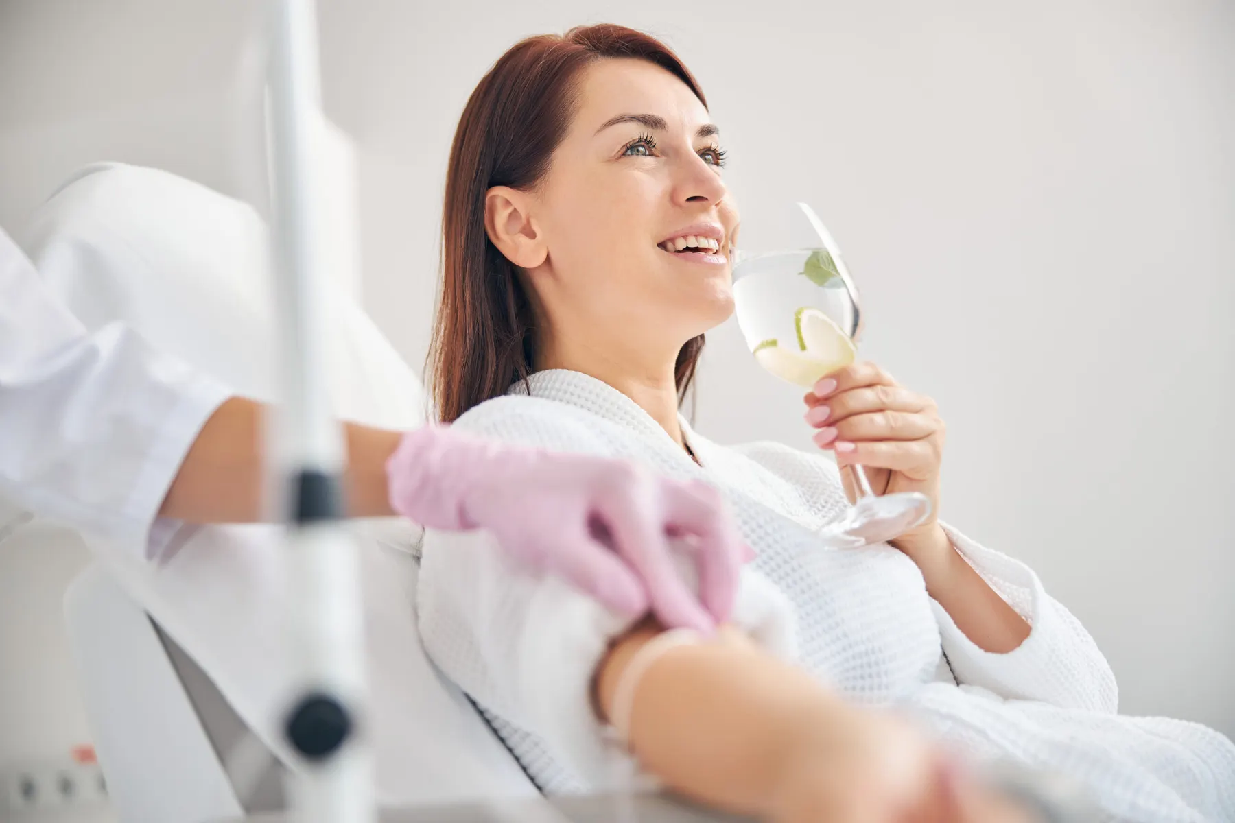 A client reclining in a treatment chair in a robe, holding a drink while a practitioner in pink gloves works on her arm.