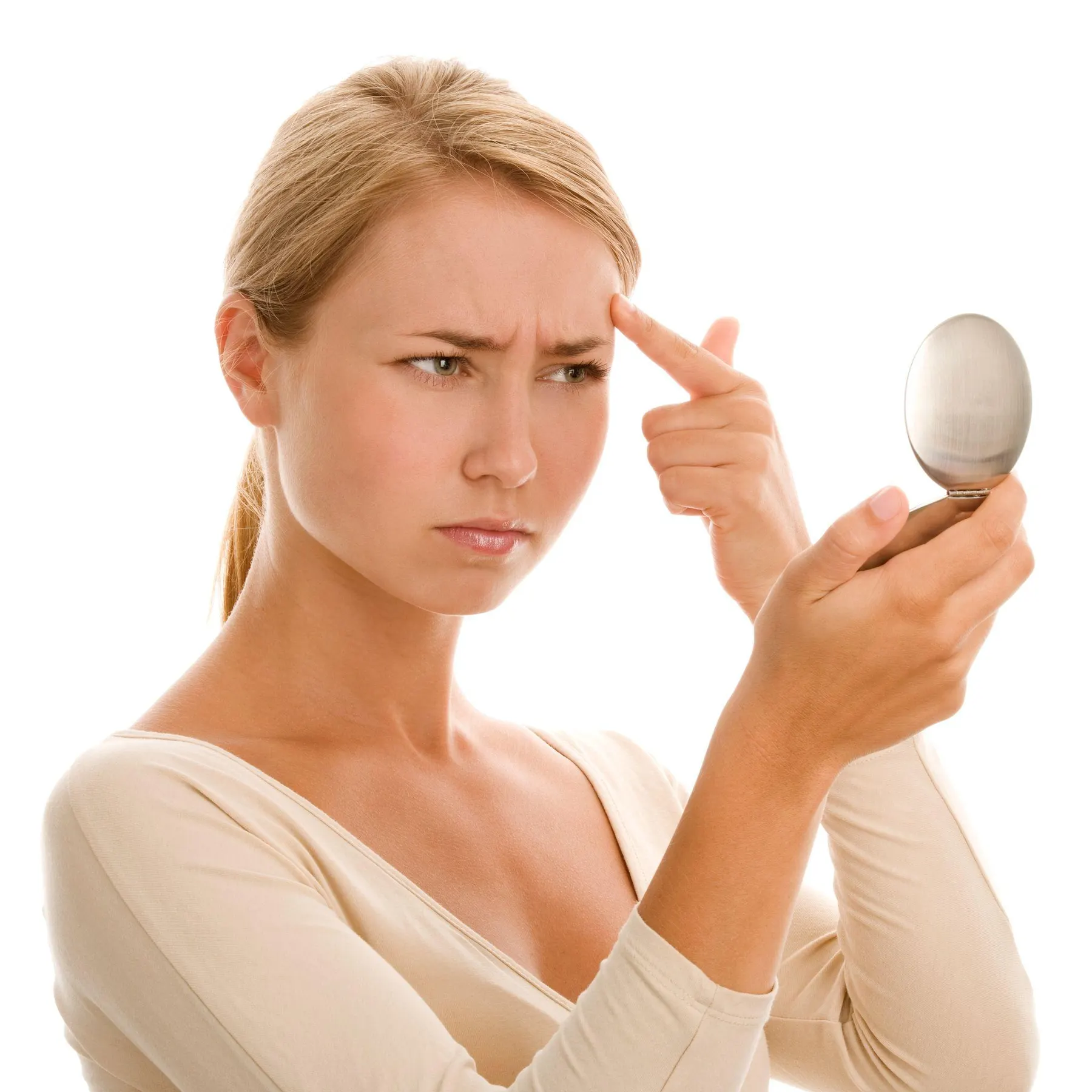 A blonde woman frowns into a small handheld compact mirror while pressing a fingertip against a line above her eyebrow.