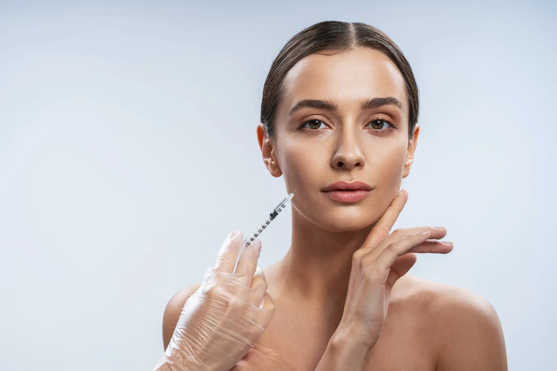 A woman resting her hand under her chin while a gloved hand places a fine syringe against her cheek.