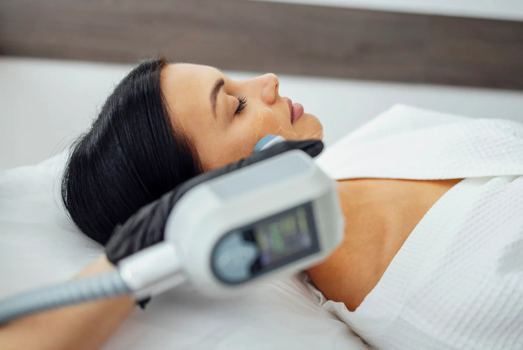 A treatment handpiece held against the cheek of a client lying back on a clinic bed.