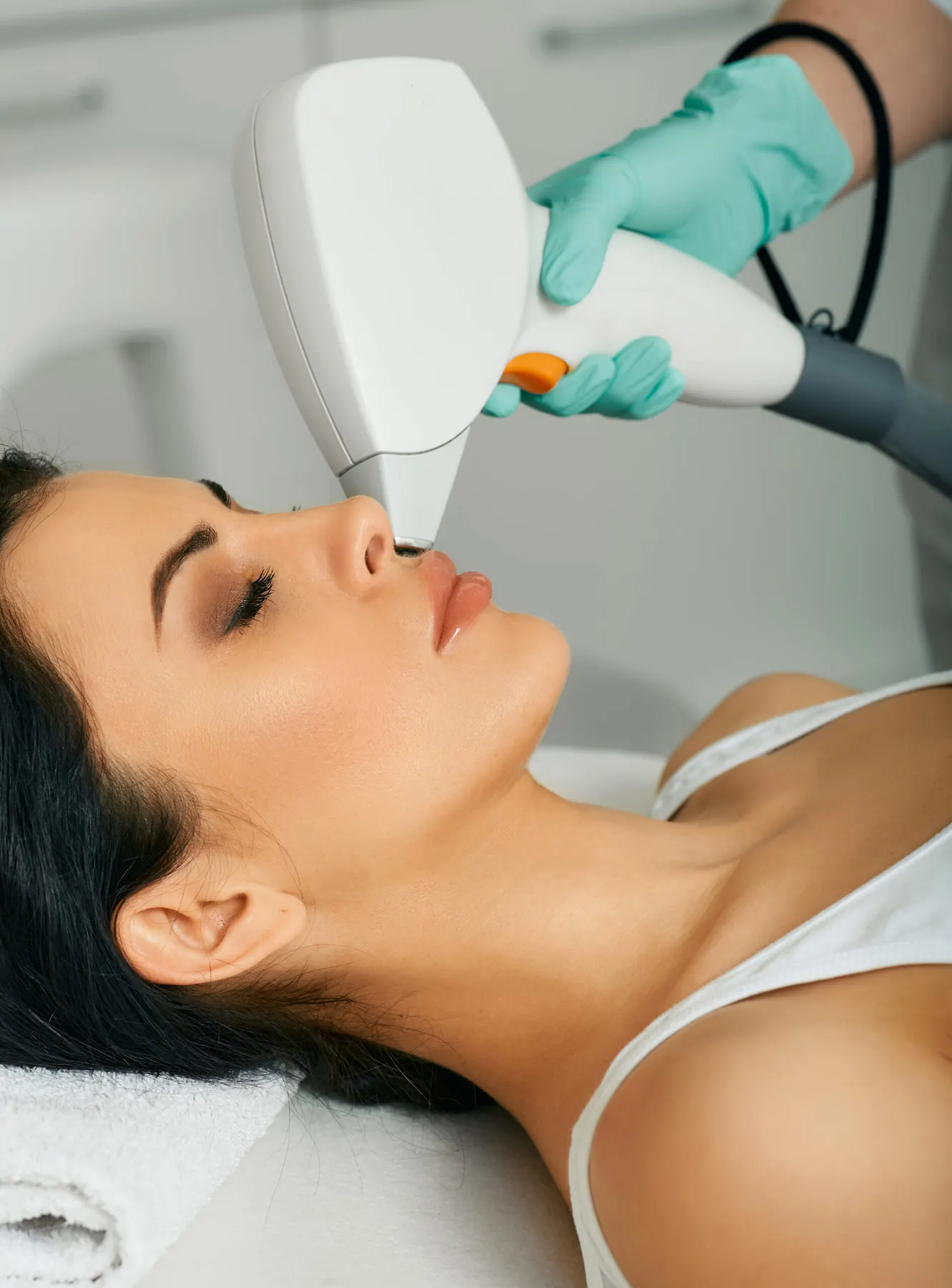 A practitioner in teal gloves holding a laser handpiece against the jaw of a client lying on a clinic bed.