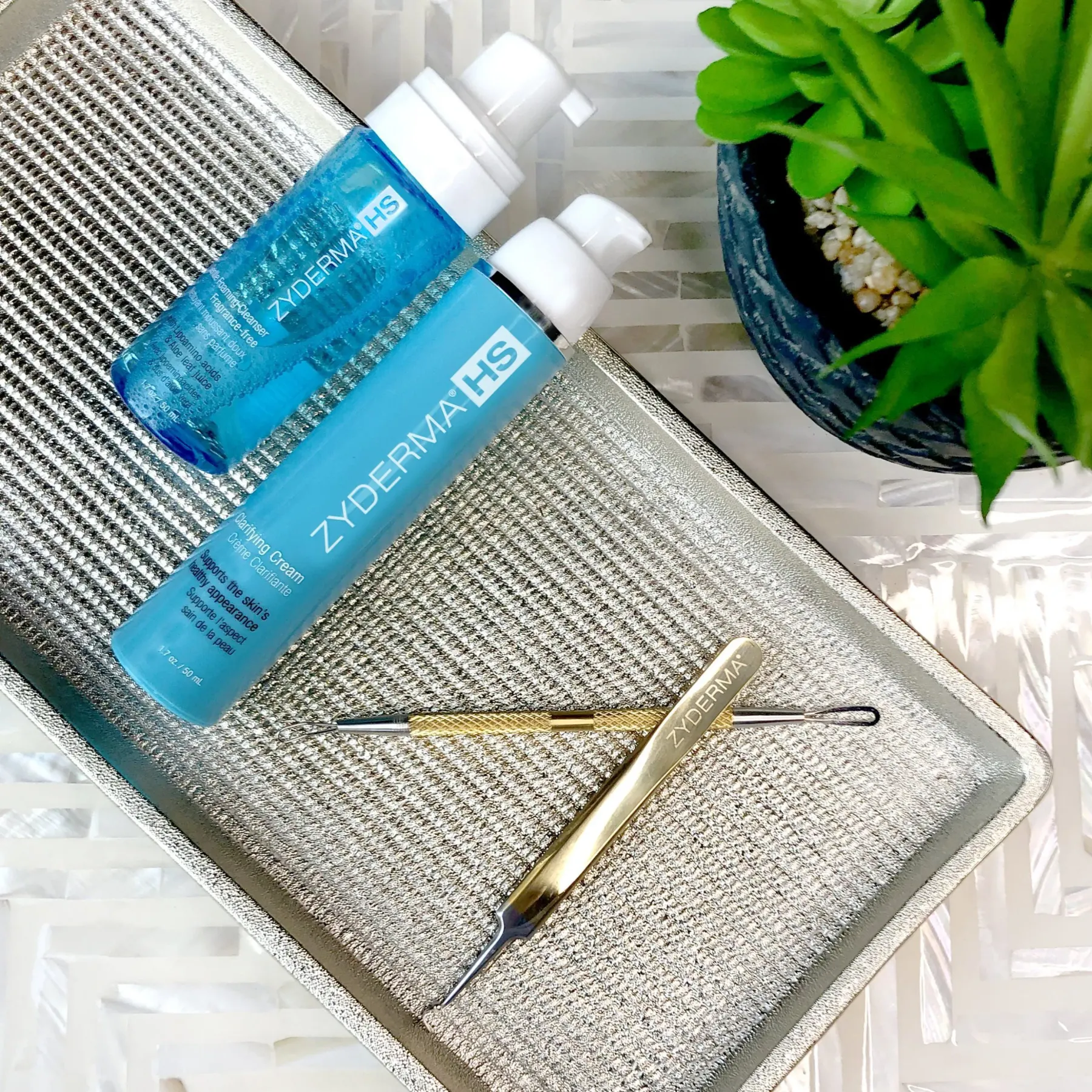 A prepared clinic tray holding two bottles of skincare, a small plant and two stainless steel extraction tools.