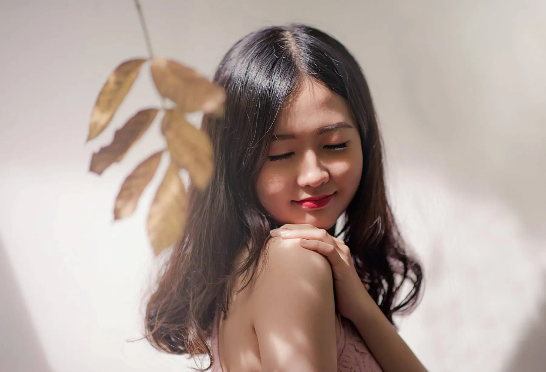 A woman with her eyes closed and her hand at her shoulder, a dried leaf held beside her in warm light