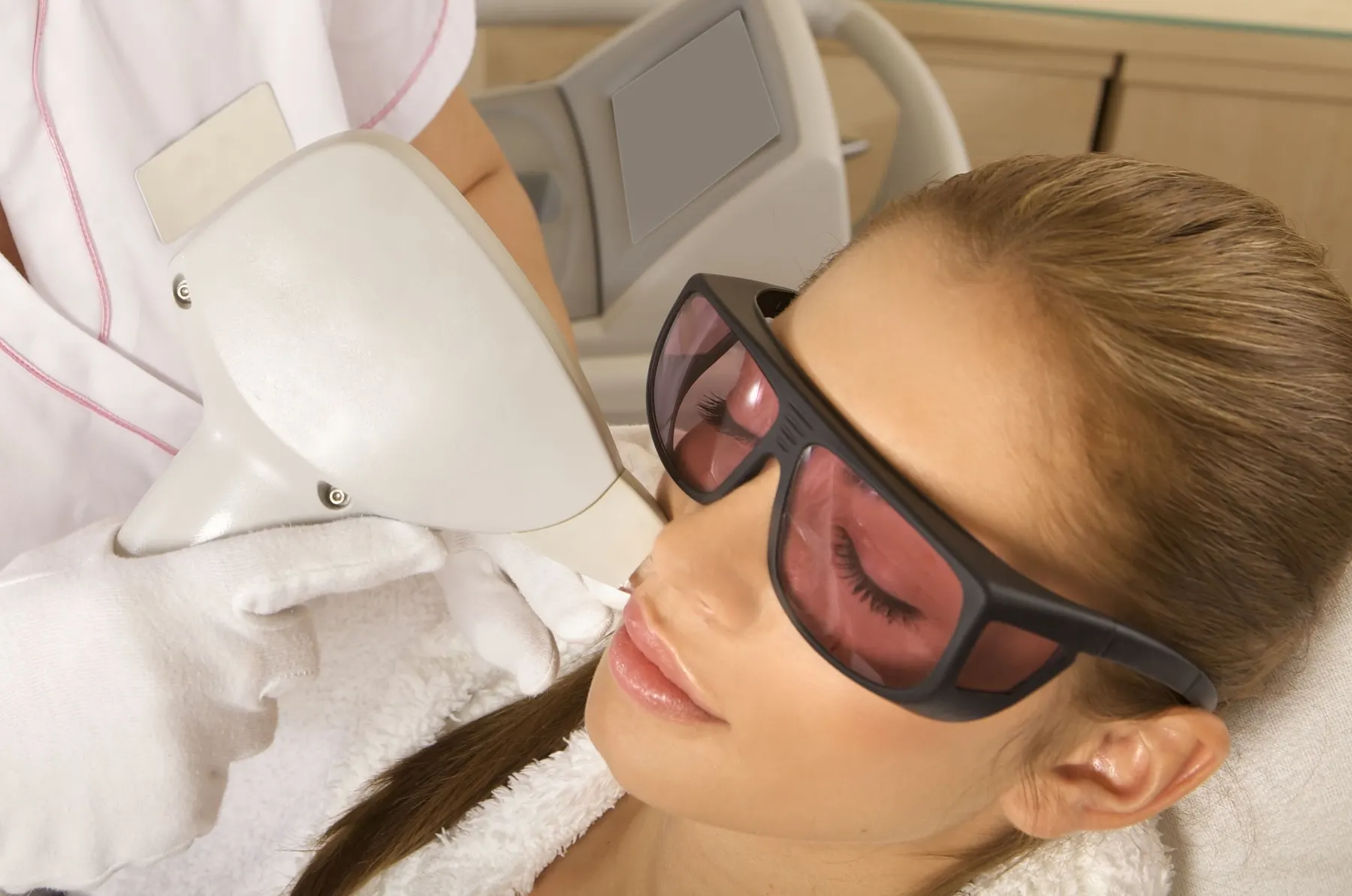 A client wearing pink-tinted protective goggles while a laser handpiece is held over her face