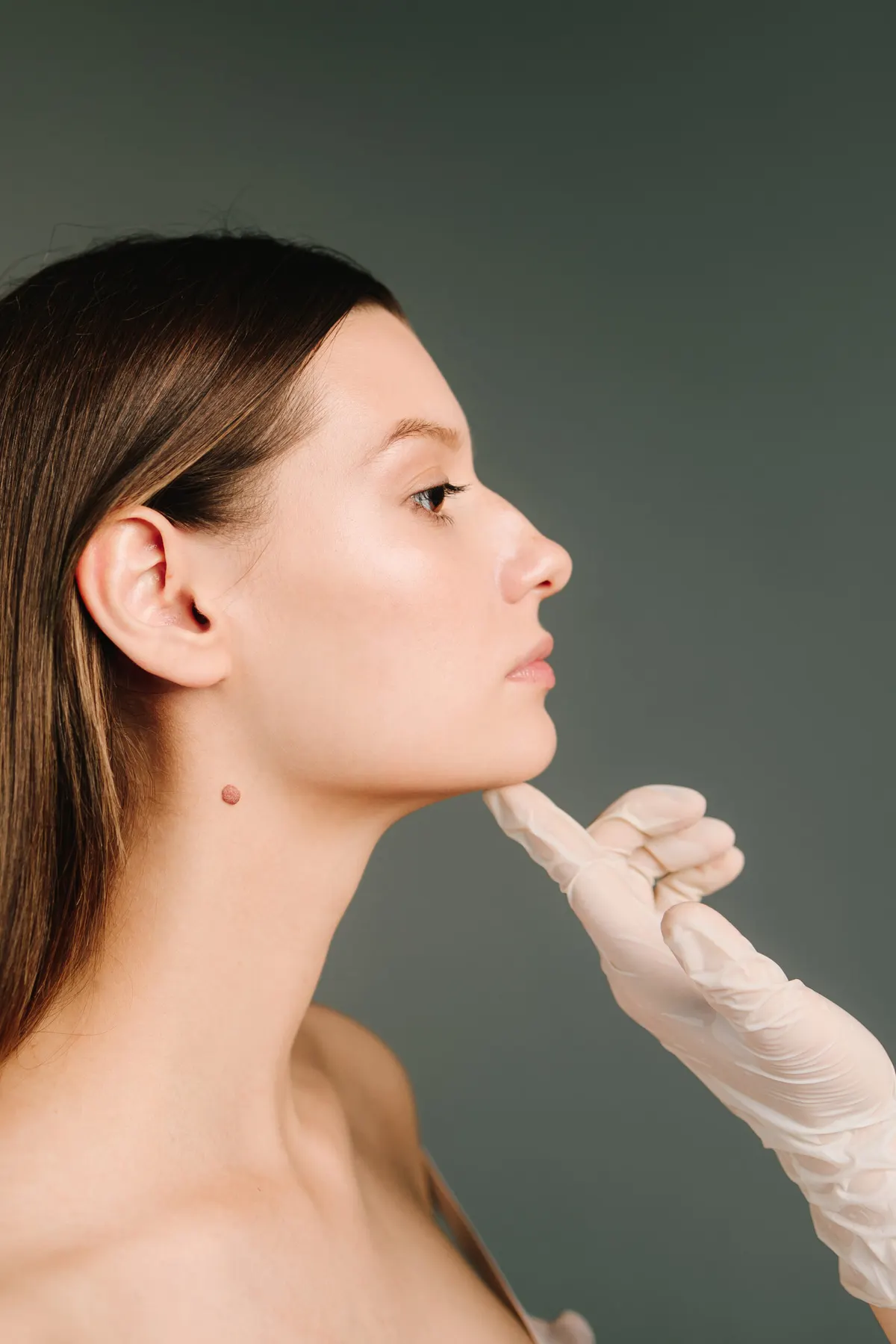 A gloved hand tilting the chin of a client upward so the side of her face can be examined in profile.