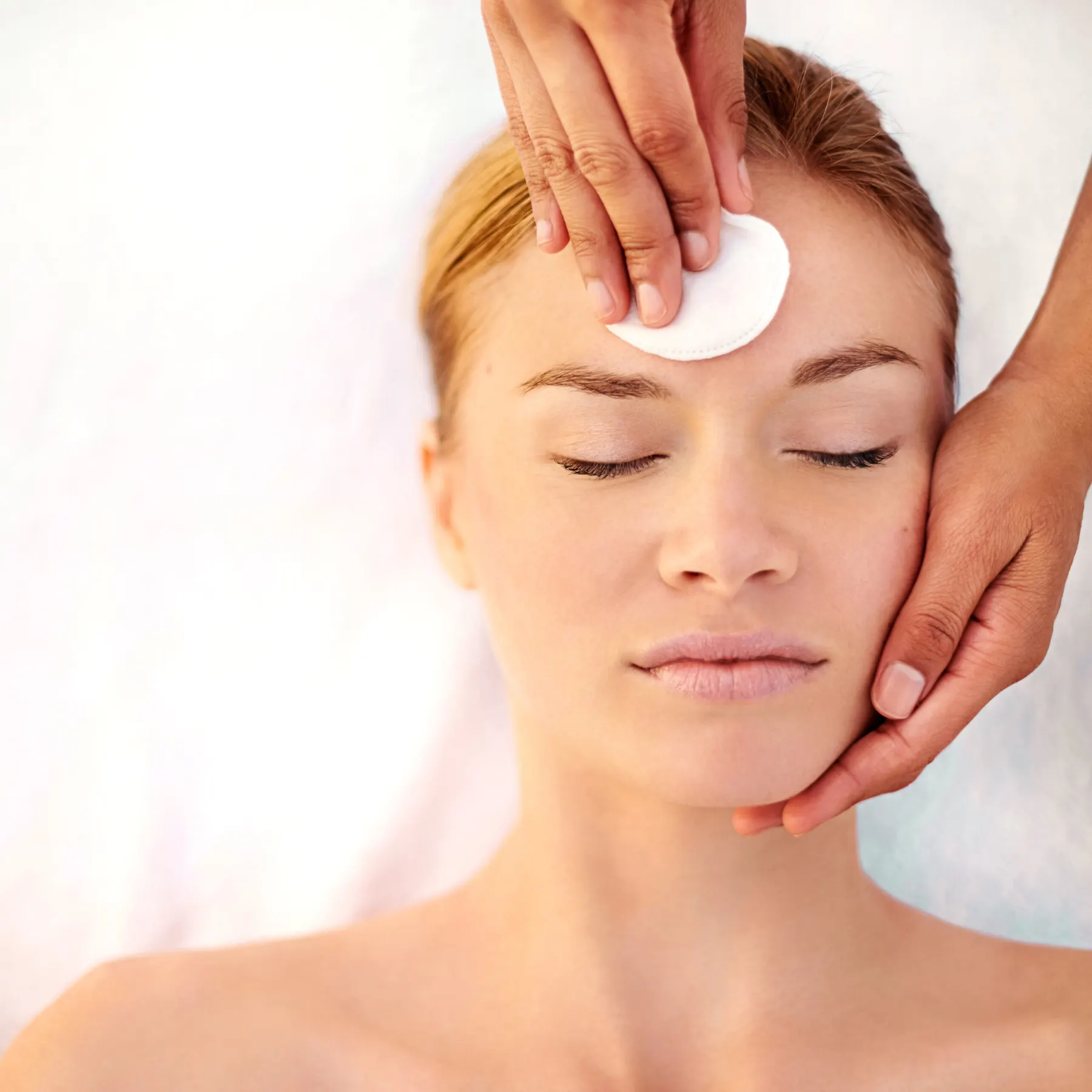 A practitioner sweeping a cotton pad across the forehead of a client who is lying back with her eyes closed.