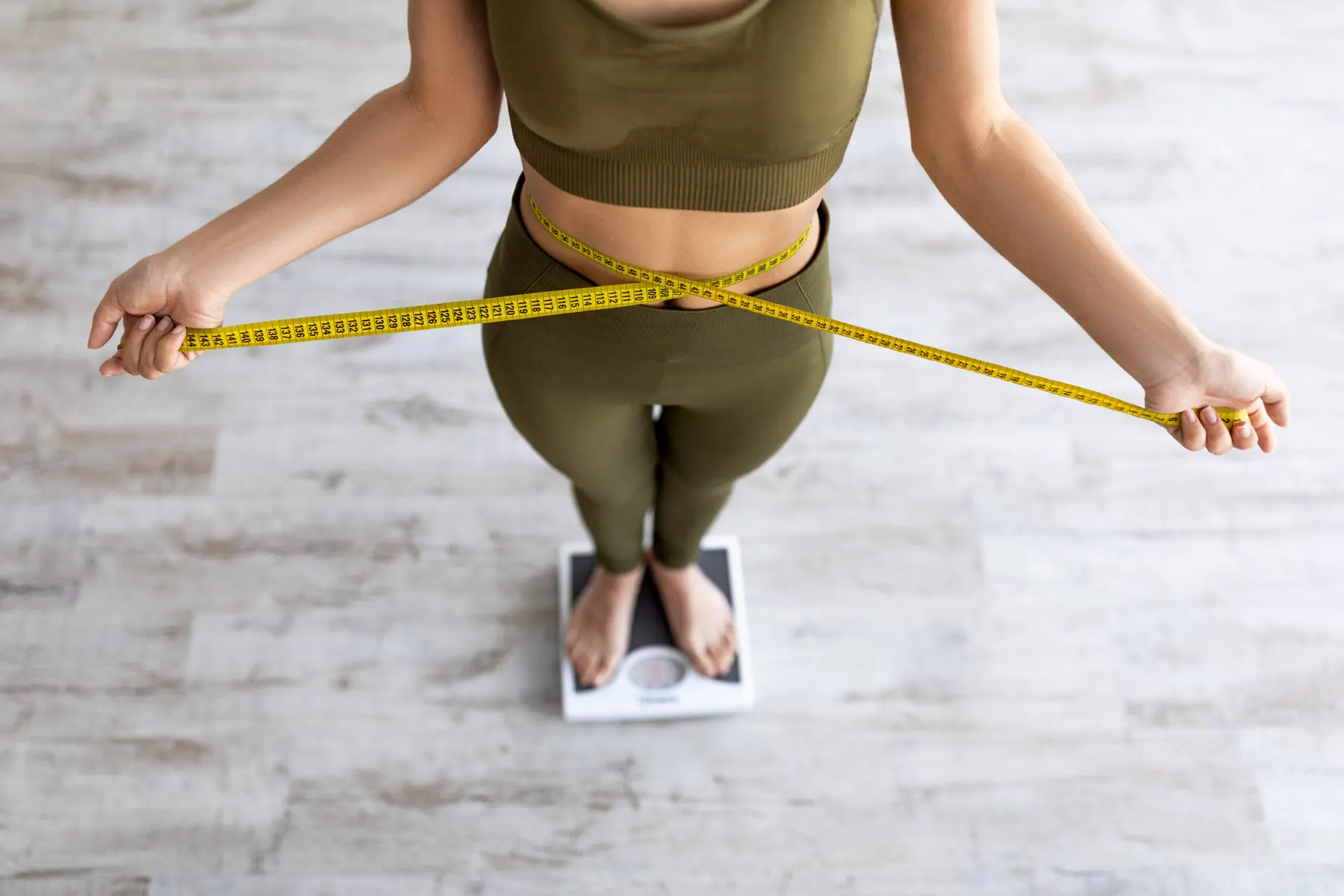 A woman in workout clothes measuring her waist with a tape measure in a bright room.